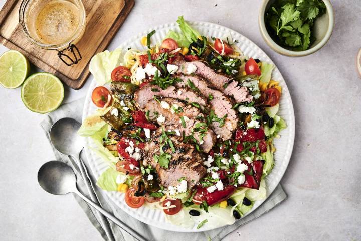 A vibrant salad topped with sliced grilled beef, assorted veggies, feta cheese, and cilantro, accompanied by lime halves and dressing.